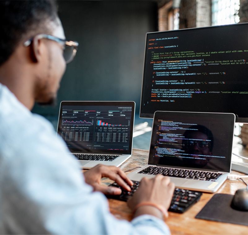 An AWS Solutions Architect sits at a desk with three screens displaying code, dashboards, and cloud-infrastructure visuals, designing and optimizing cloud architectures in a modern workspace.