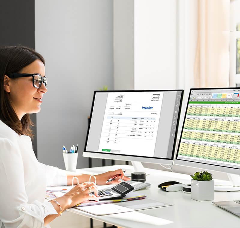 A smiling woman wearing glasses sits at a modern white desk, working on medical coding and billing tasks. She uses a calculator and reviews paperwork in front of two computer monitors displaying spreadsheets and invoice data.
